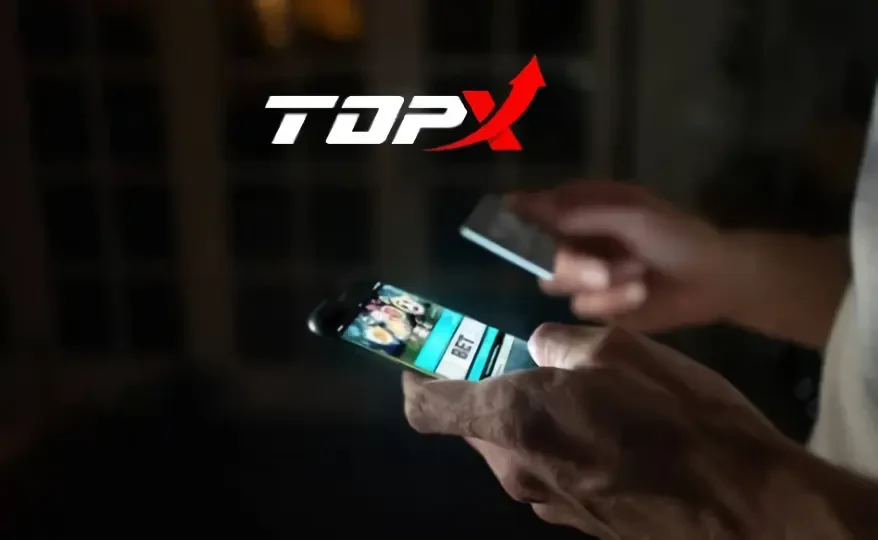 Mãos segurando um celular no escuro, com o logotipo da TopX ao fundo
